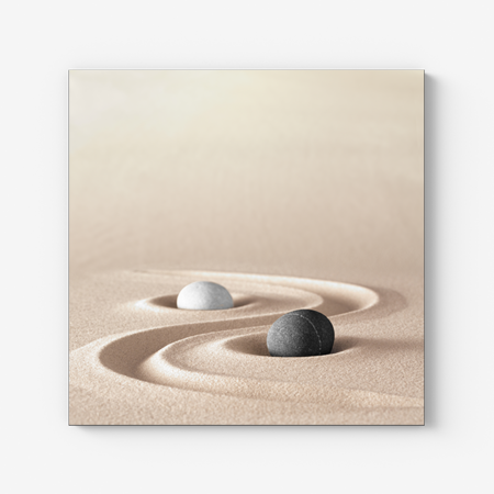 Das Bild zeigt einen schwarzen und weißen Stein auf dem Sand liegend. Dazwischen wurde eine gewellte Linie in den Sand gezeichnet und formt ein beruhigendes Yin und Yang Symbol.