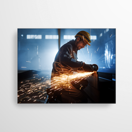 Das Bild zeigt ein Akustikpaneel mit einem Industrie Motiv. Der Arbeiter trägt einen Schutzhelm und schneidet ein Metallrohr, sodass Funken sprühen. Das Bild zeigt den normalen Produktionsalltag in der Industrie.