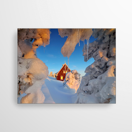 Dieses Akustikbild zeigt den Blick in ein Winterwunderland. Die schneebedeckten Äste eines Baumes rahmen das Bild ein. Man scheint durch den Schnee hindurch zu gucken und sieht eine Holzhütte, die in das warme Abendlicht der Sonne getaucht wird.